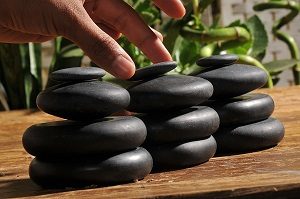 Trois piles de quatre pierres de massage noires. Les pierres sont joliment disposées sur une table en bois avec des plantes vertes au fond. Une main s’approche. 